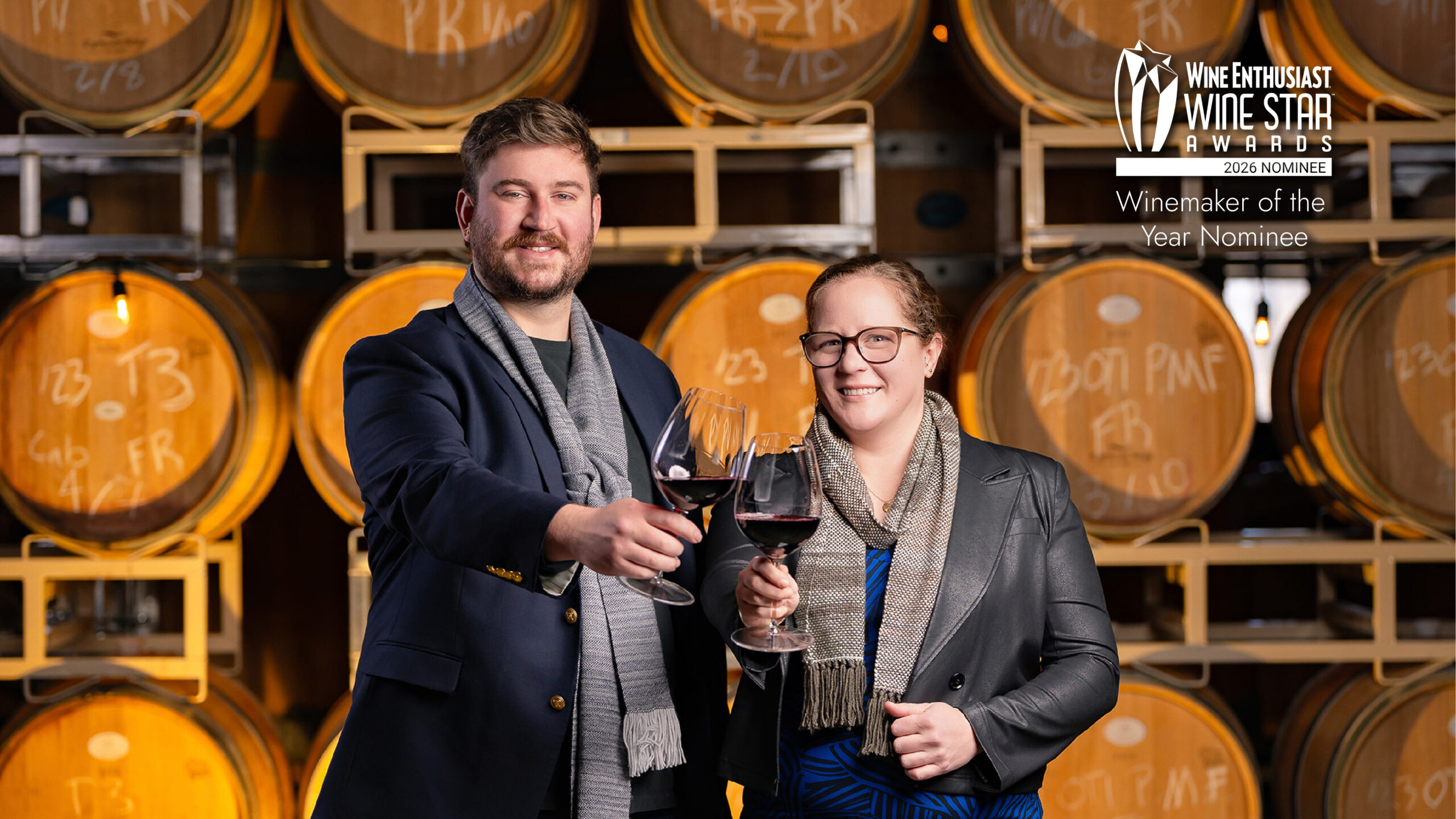 Two people cheering glasses of red wine with barrels in the background.